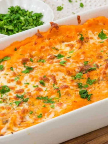 A baked sweet potato casserole topped with melted cheese and chopped parsley, served in a white rectangular dish on a wooden board. Small bowls of fresh herbs and seasonings are visible in the background.