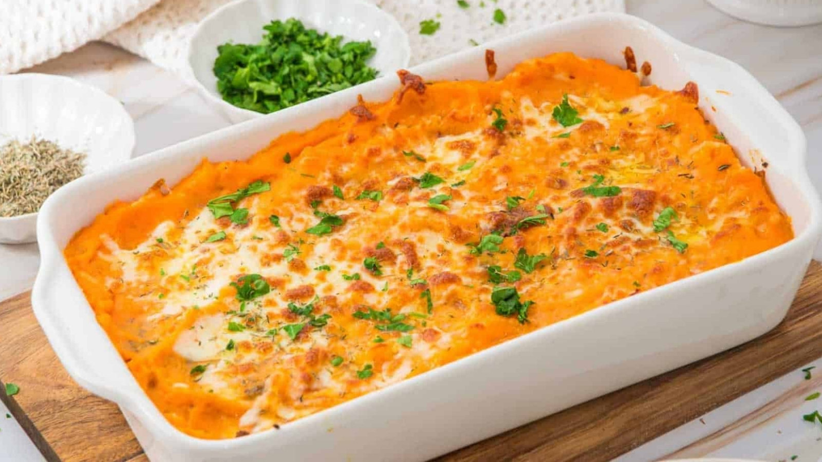 A baked sweet potato casserole topped with melted cheese and chopped parsley, served in a white rectangular dish on a wooden board. Small bowls of fresh herbs and seasonings are visible in the background.