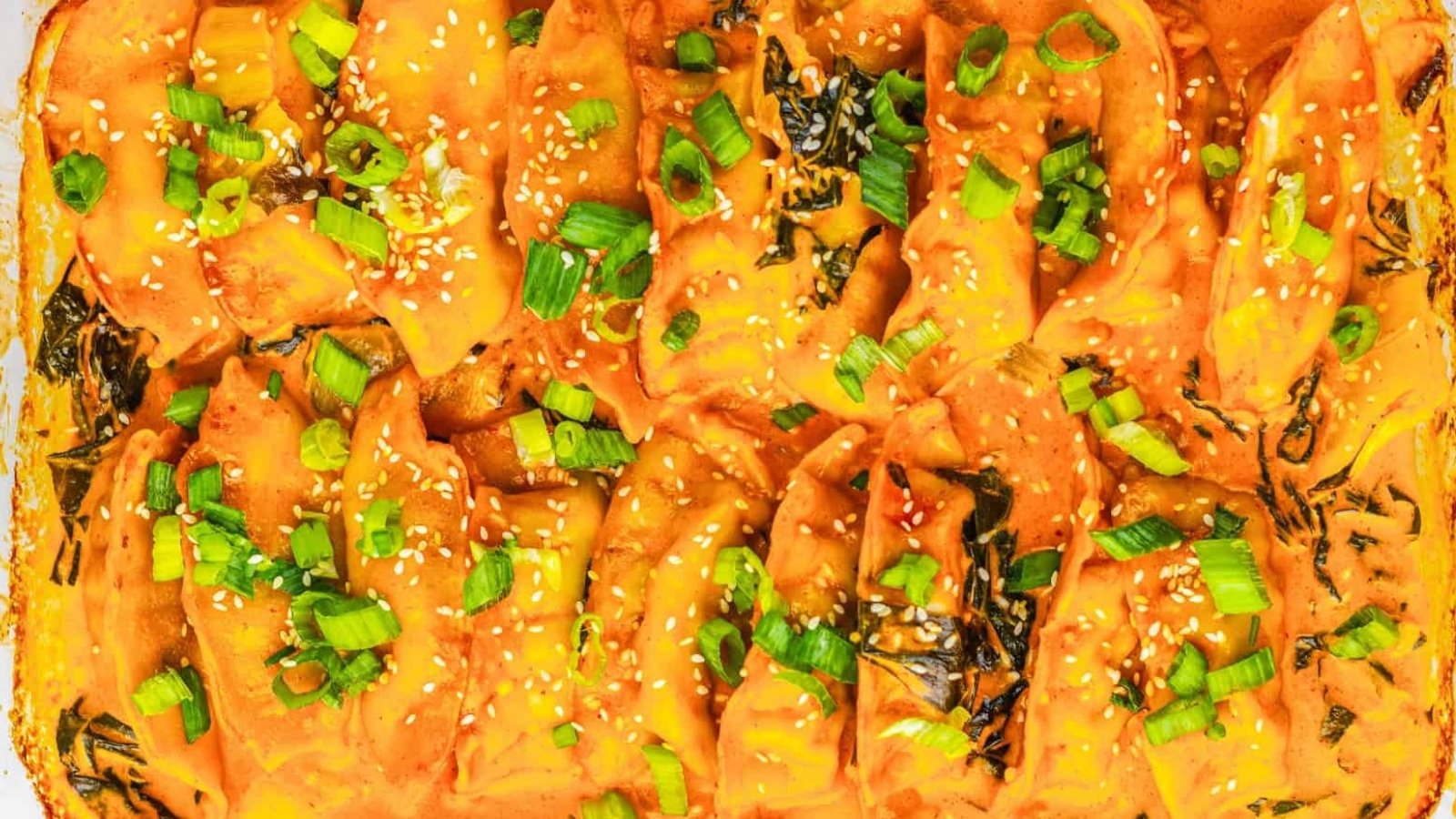 Close-up of baked dumplings in orange sauce, topped with chopped green onions and sesame seeds.