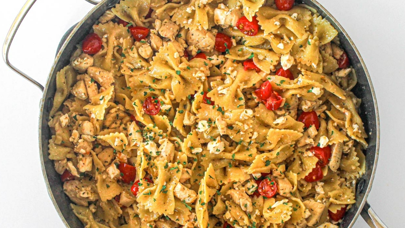 A pan filled with bowtie pasta mixed with diced chicken, cherry tomatoes, feta cheese, and chopped herbs.
