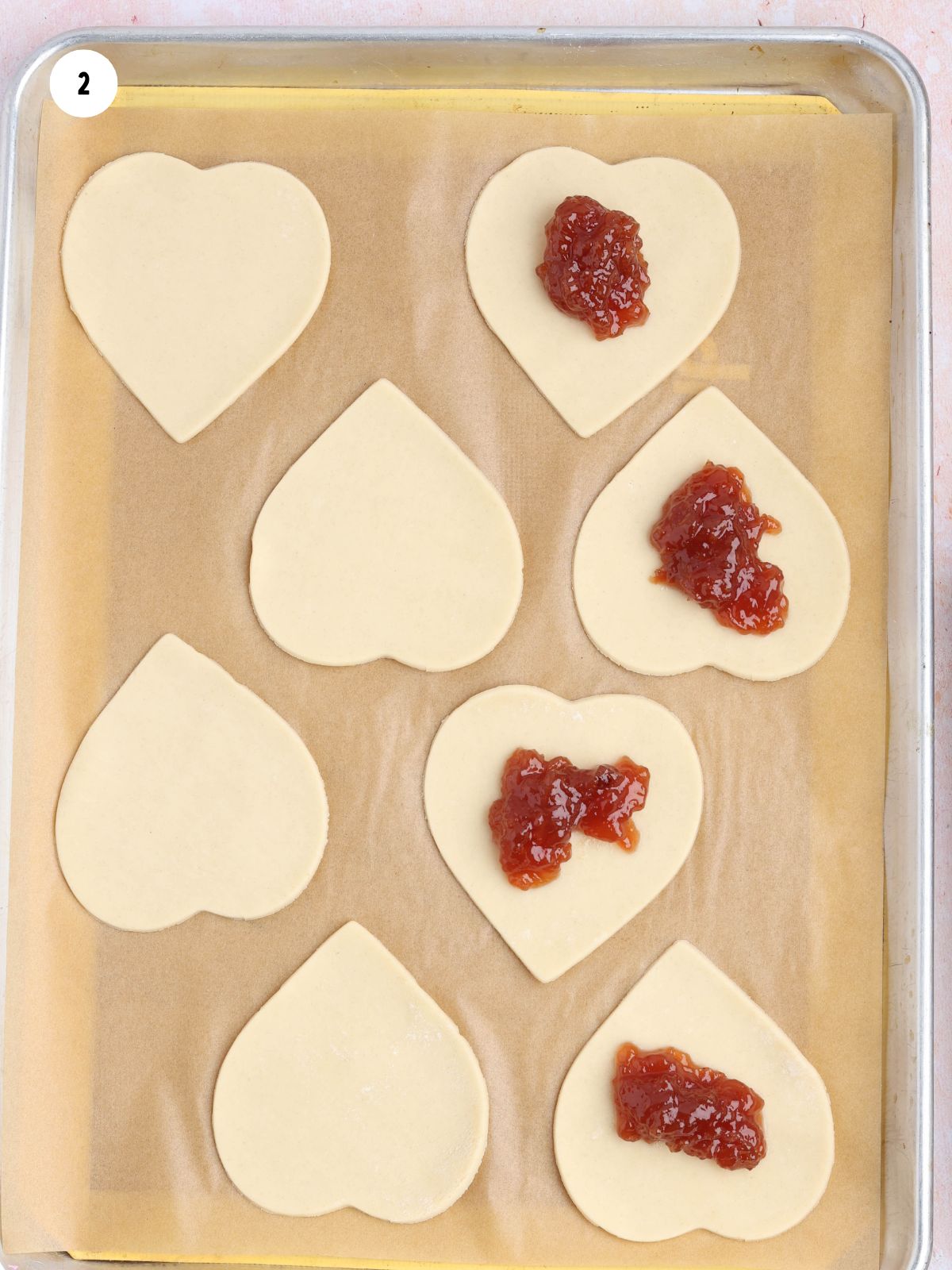 Strawberry jam spooned onto heart-shaped pie dough cutouts on a parchment-lined baking sheet.