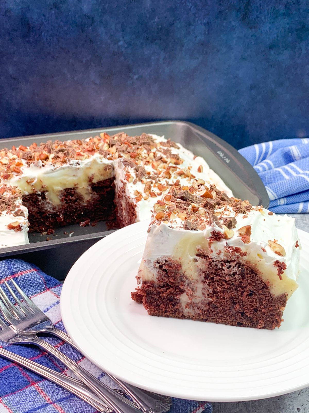 cheesecake pudding poke cake with chocolate cake mix in pan and sliced on white plate with forks.