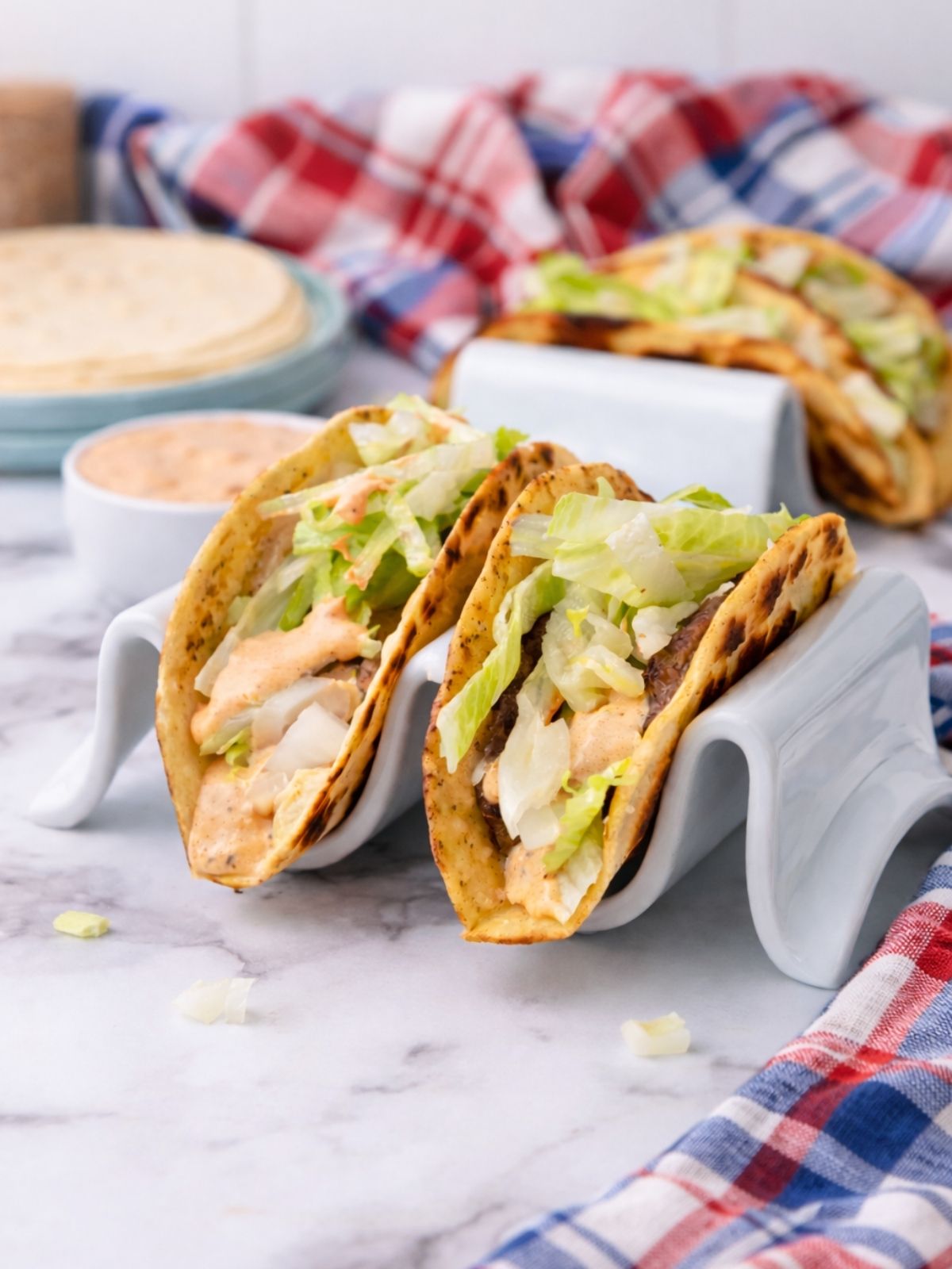 Cast Iron Smash Burger Tacos in a taco holder. Red white and blue checked towel in the background with more tacos.