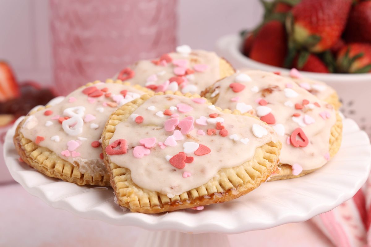Glazed heart shaped strawberry hand pies with pink and white sprinkles displayed on a white cake stand.