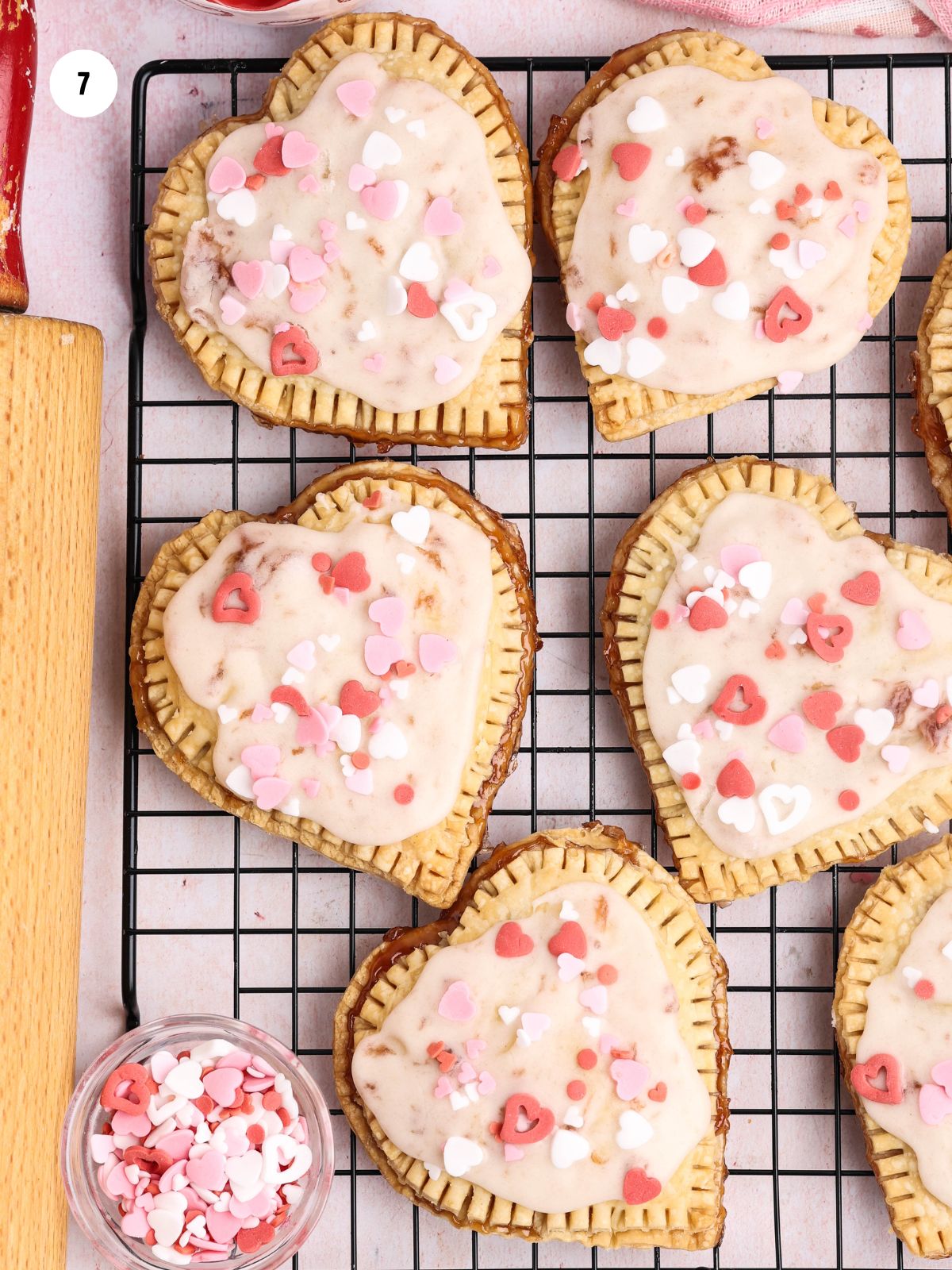 Glazed strawberry hand pies on baking rack decorated with pink and red heart sprinkles in small bowl.