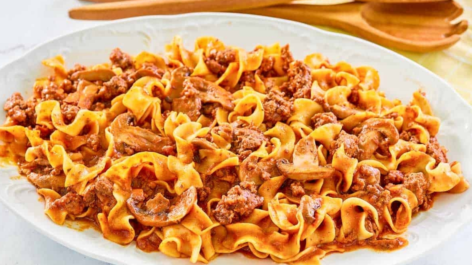 Egg noodles mixed with ground meat and sliced mushrooms in a red sauce are served on a white plate with wooden spoons in the background.