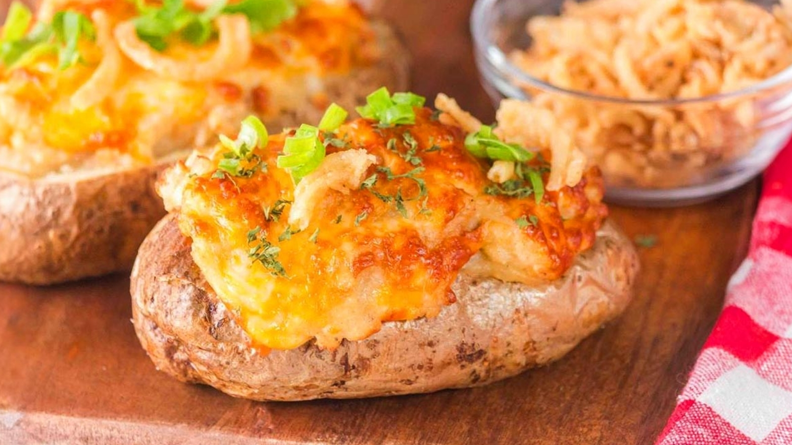 A baked potato topped with melted cheese, green onions, and crispy fried onions, with a bowl of fried onions in the background.