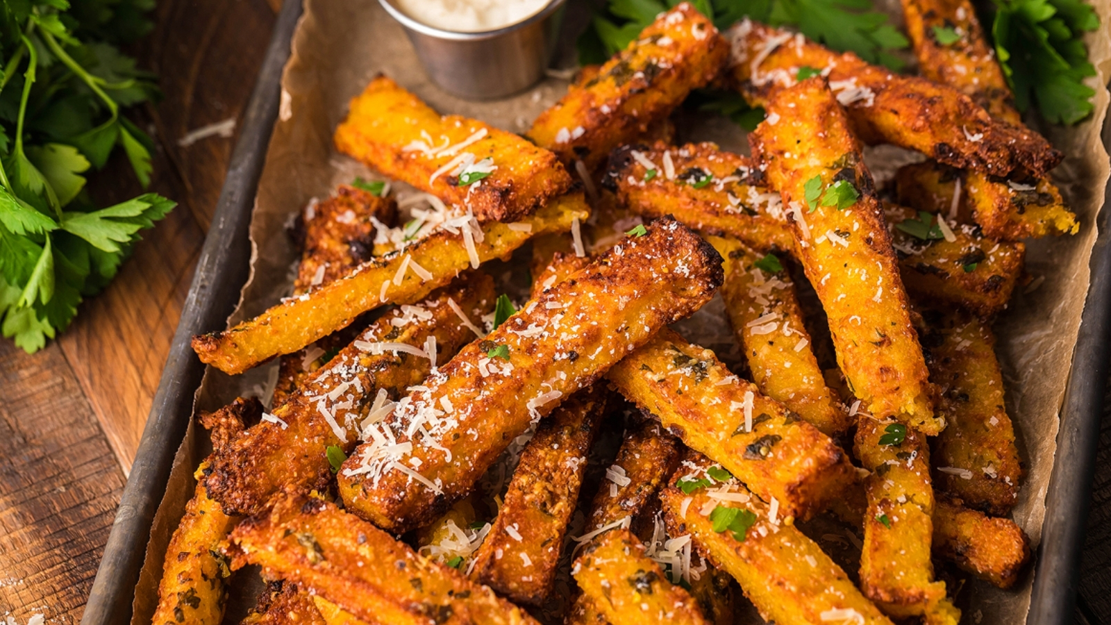 Golden polenta fries topped with grated cheese and herbs are arranged on a paper-lined tray with a small metal cup of dipping sauce and parsley beside them.