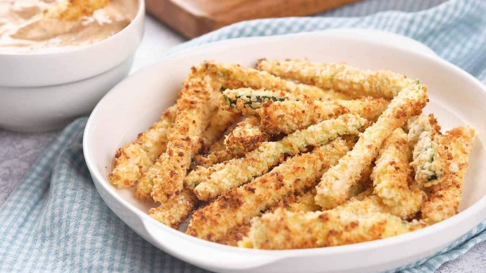 A white bowl filled with crispy, breaded air fryer zucchini fries is placed on a blue and white cloth.