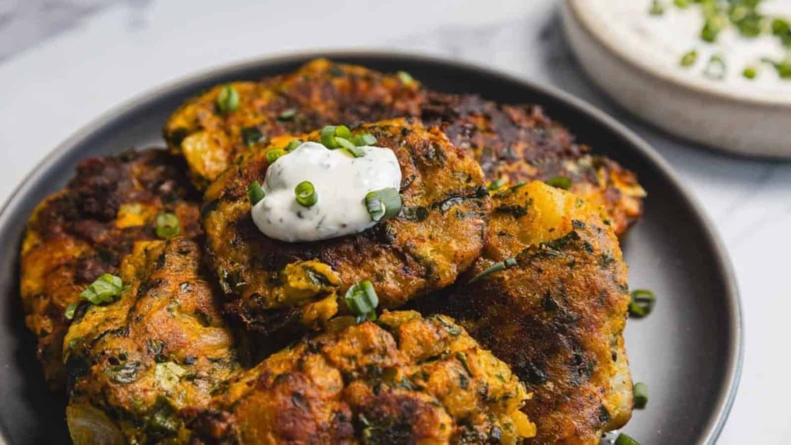 A plate of golden-brown fritters topped with creamy dip and chopped green onions, with more dip in the background.