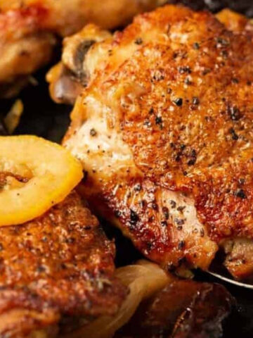 Crispy roasted chicken thighs in a pan with sliced apples, onions, and dark dried fruit, with a spoon lifting one piece.