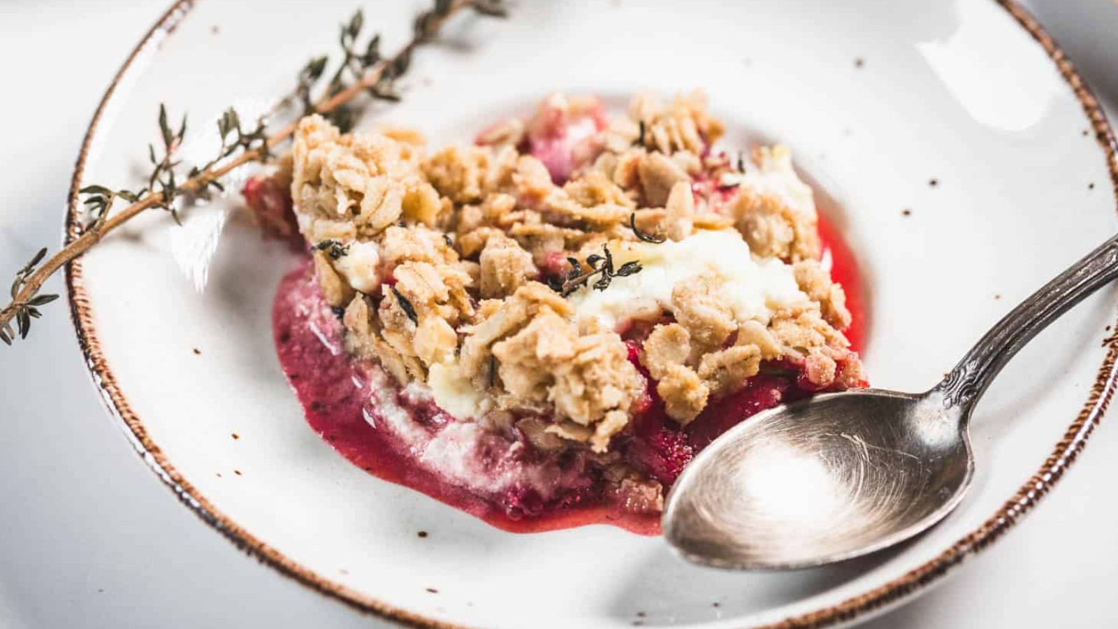 A serving of fruit crumble with a crumbly oat topping and a layer of red fruit filling, garnished with a sprig of thyme on a white plate with a brown rim, next to a silver spoon.