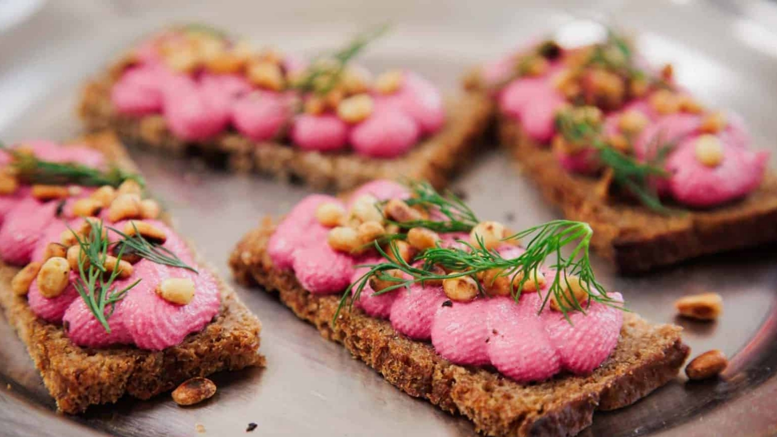 Four rectangular pieces of brown bread topped with pink spread, toasted pine nuts, and fresh dill are arranged on a metal tray.