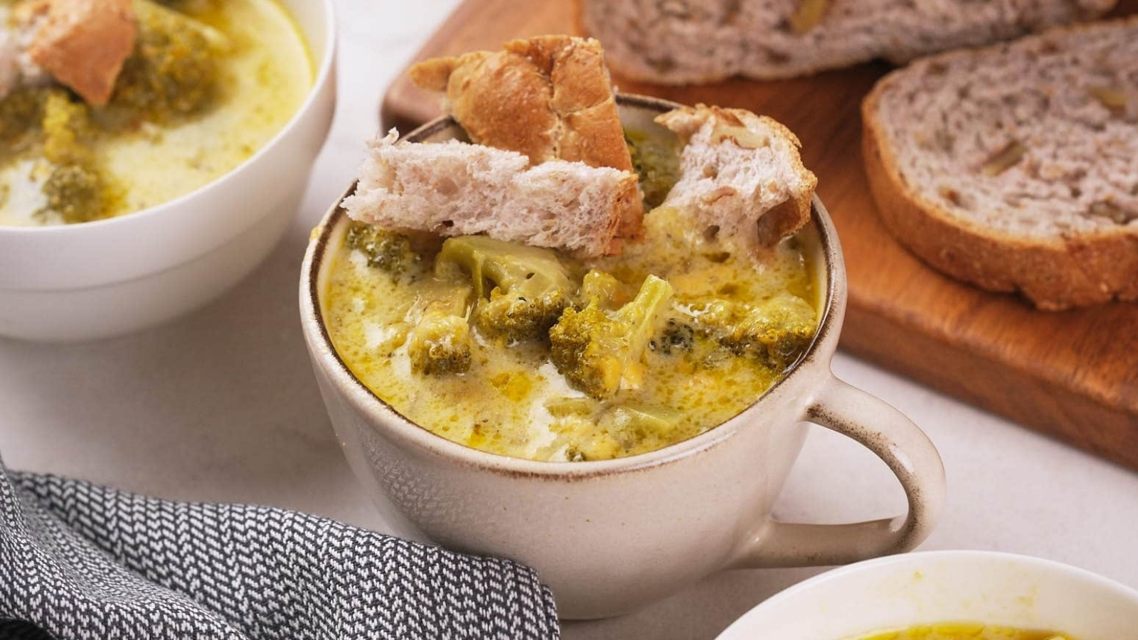 A bowl of broccoli cheddar soup topped with bread pieces next to sliced bread on a wooden board.