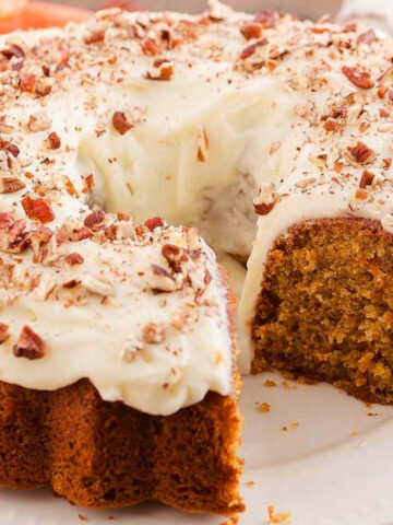 A frosted bundt cake topped with chopped nuts sits on a white plate with one slice cut out and placed slightly apart.