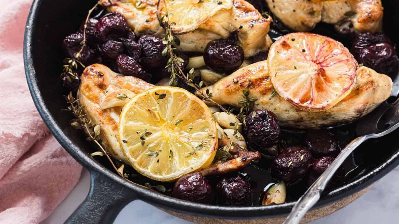 Skillet with roasted chicken breasts, lemon slices, grapes, herbs, and garlic cloves.