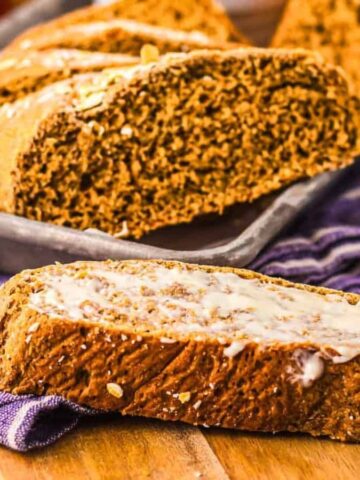 A slice of brown bread with butter sits in front of a tray of sliced bread on a purple cloth.