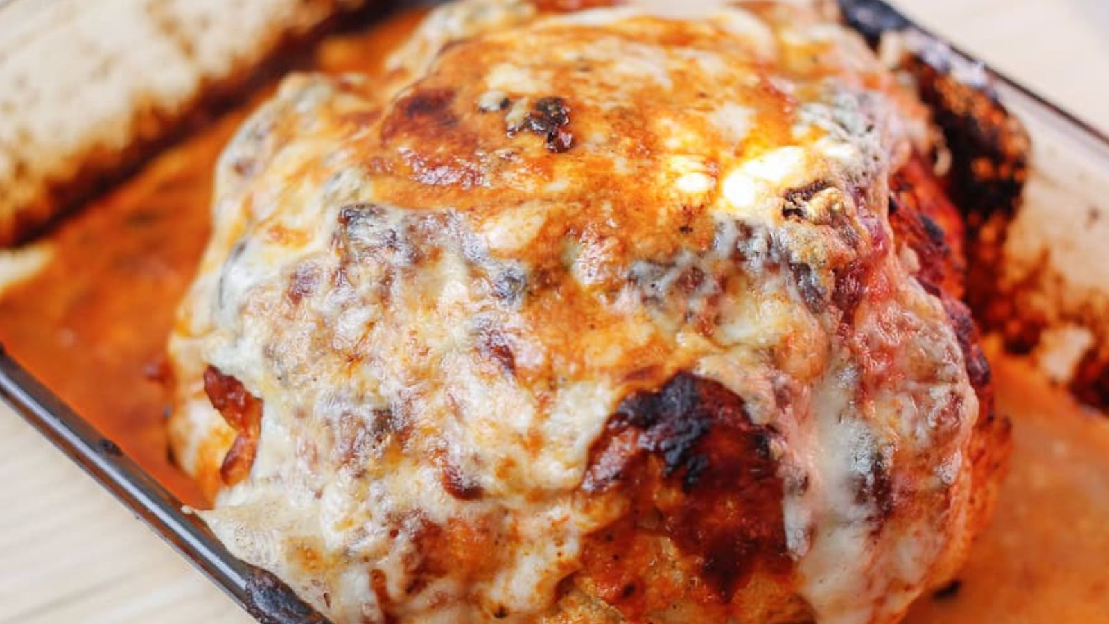 A baked dish with a large serving of cheesy cauliflower in a glass baking dish is next to a white plate holding a portion of the same dish. The background is a light wooden surface. A fork is visible beside the plate.