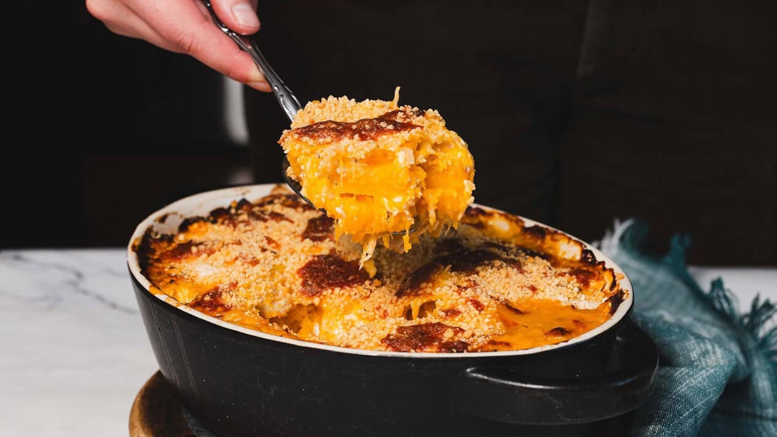 A person lifts a spoonful of cooked macaroni and cheese from an oval black baking dish. The dish is topped with a breadcrumb crust, and a blue cloth is partially visible underneath. The table surface is white.