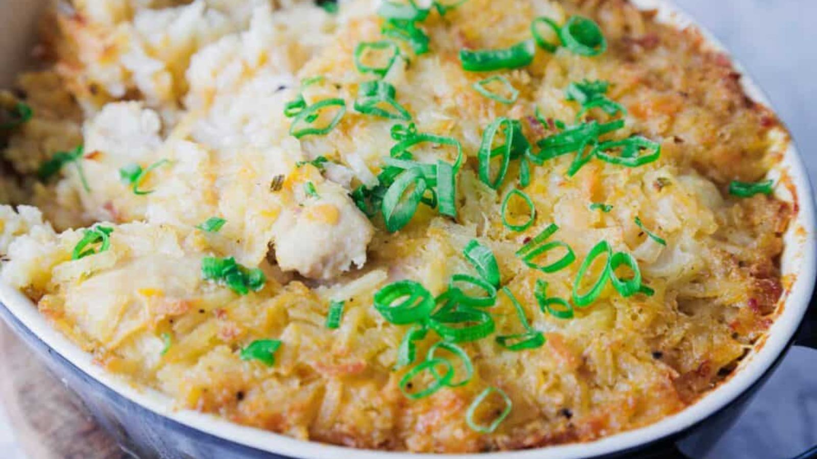 A dish in a black baking pan filled with baked casserole. The top is golden brown with crispy edges and garnished with sliced green onions. A portion has been removed, revealing a creamy interior.