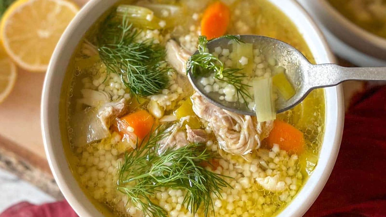 A bowl of chicken pastina soup sits on a rustic wooden cutting board with lemons and dill in the background.