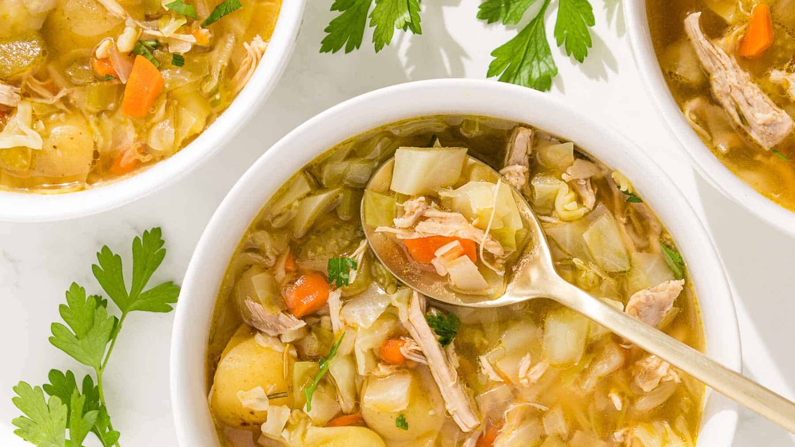 A bowl of chicken and cabbage soup is shown on a rustic wooden cutting board with parsley and a red linen.