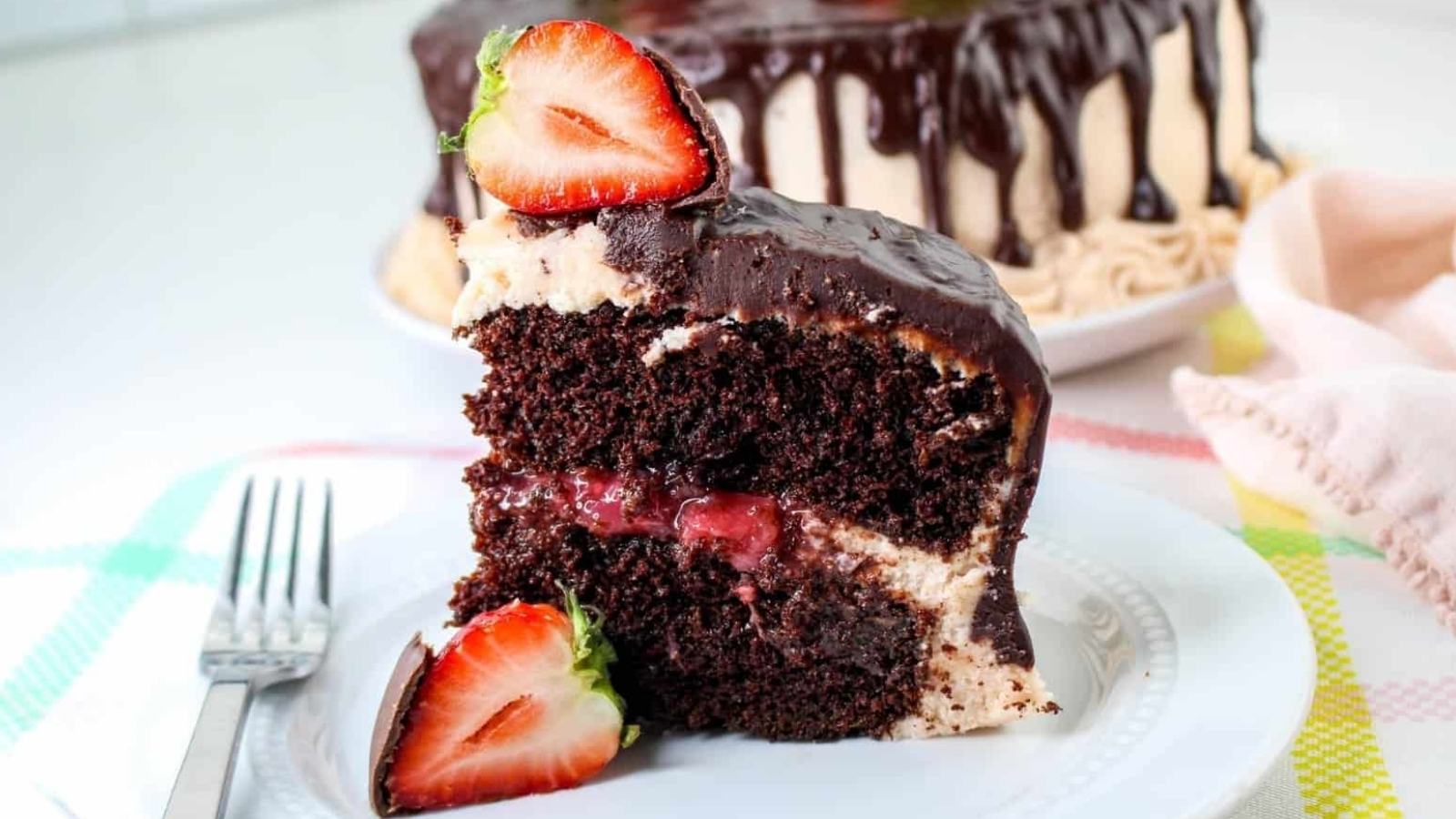 A slice of chocolate cake with a strawberry filling and dark chocolate glaze on a plate. Whole cake in the background, garnished with a halved strawberry. Fork on the side.
