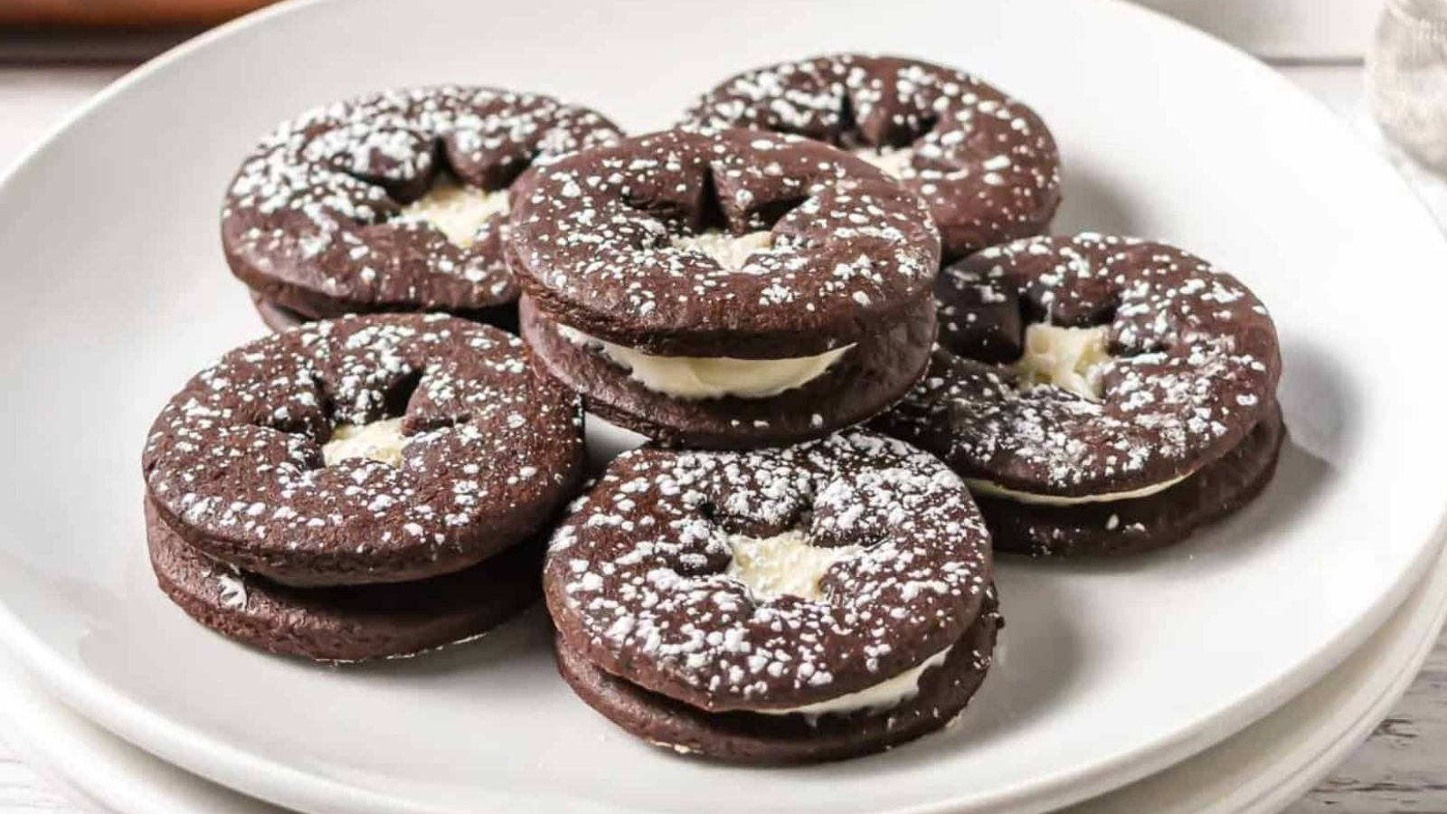 Chocolate sandwich cookies with cream filling, dusted with powdered sugar, arranged on a white plate.