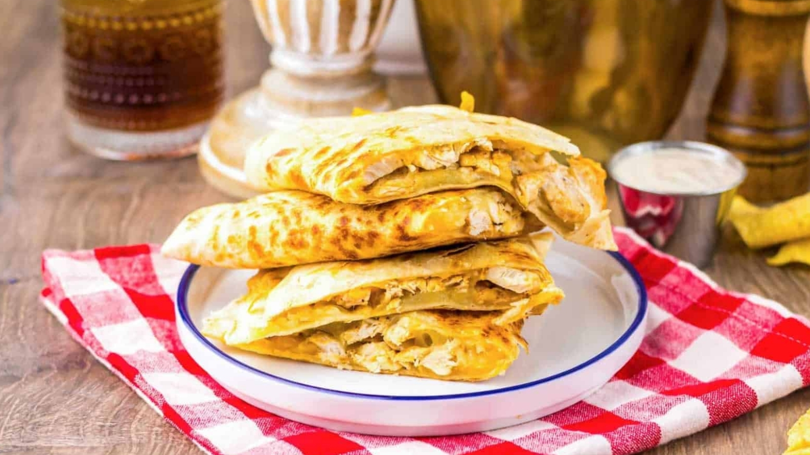 Three pieces of stuffed flatbread stacked on a plate, on a red checkered cloth, with sauce on the side.