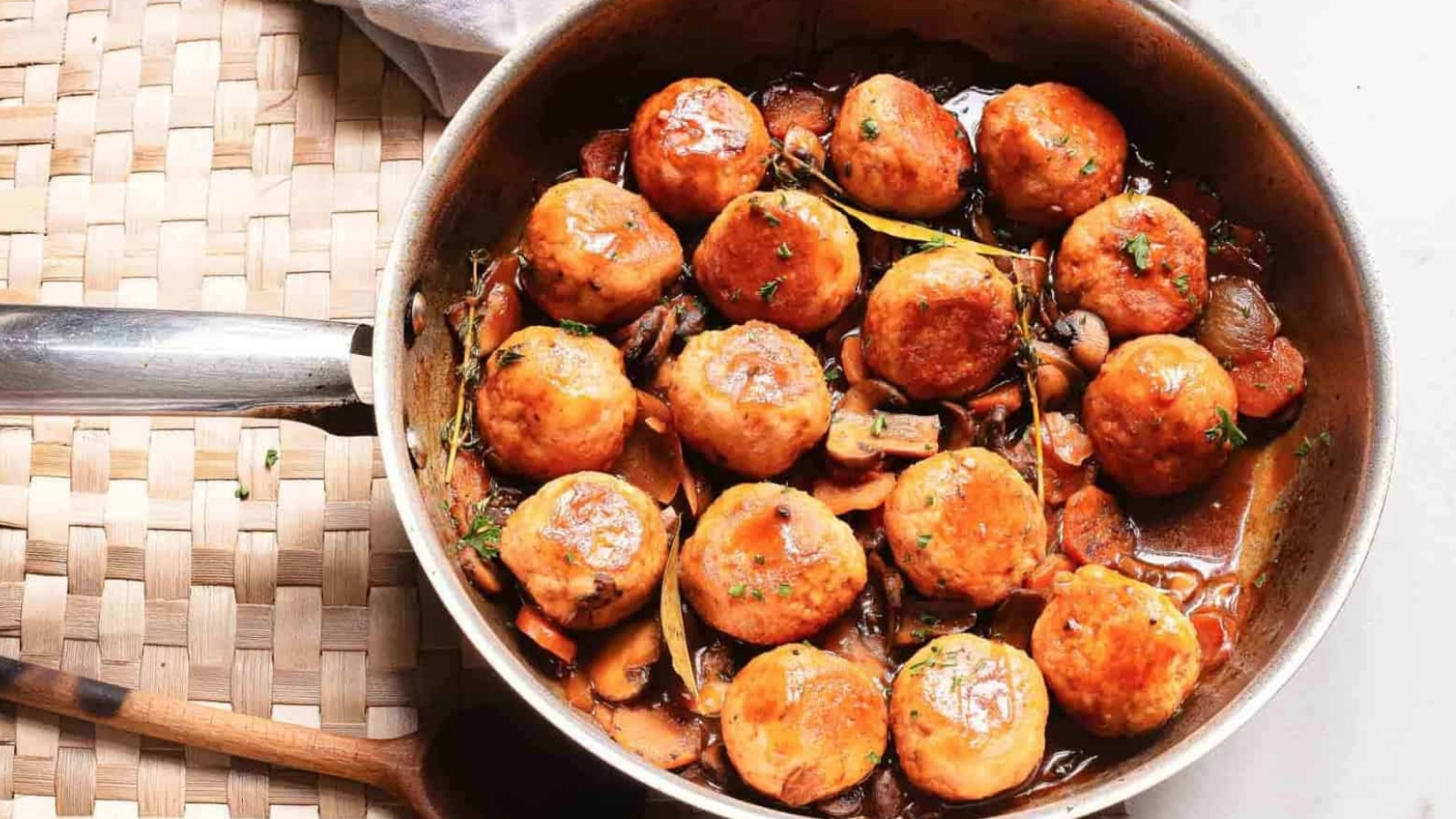 A skillet of glazed meatballs in sauce with mushrooms and herbs, viewed from above.