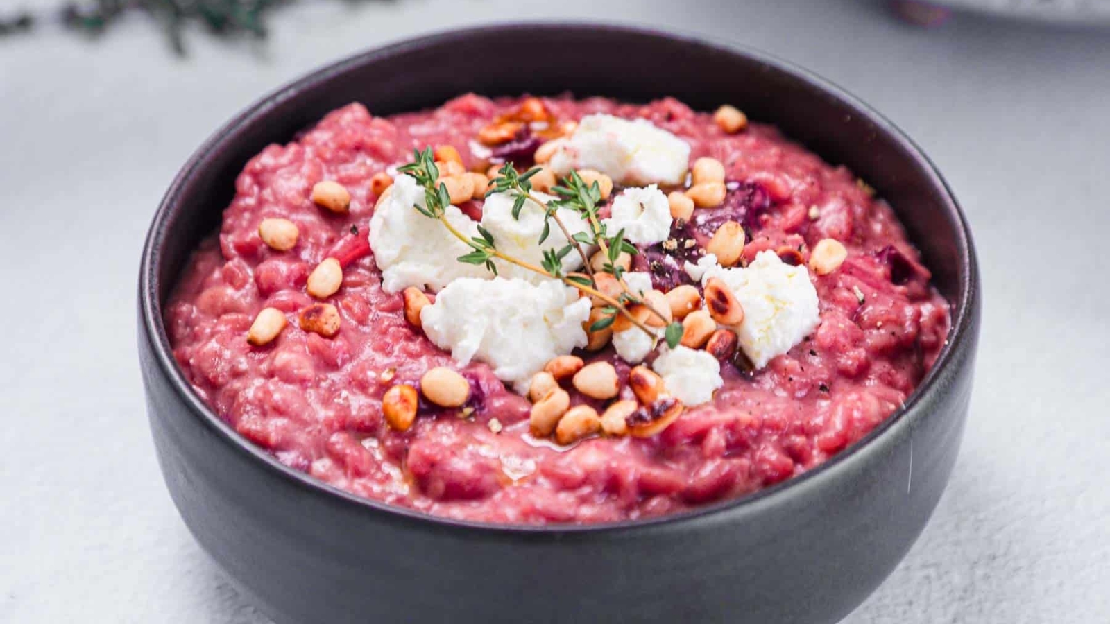 A black bowl filled with beetroot risotto, topped with pine nuts, goat cheese, and a small sprig of thyme, placed on a light-colored surface.