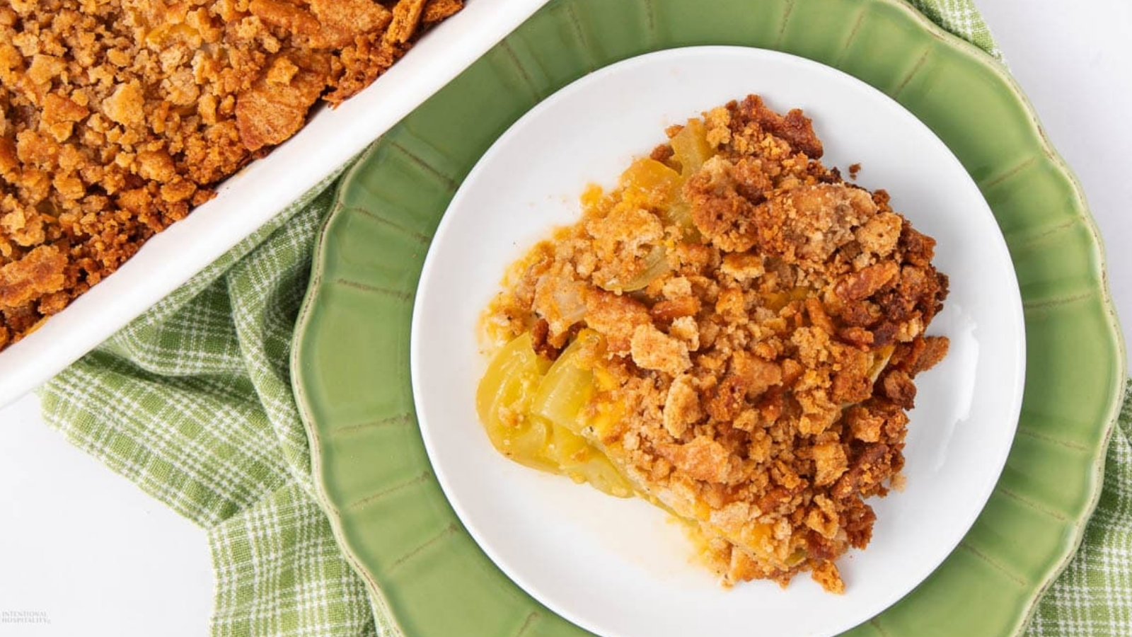 A slice of crumb-topped casserole with visible green tomato slices sits on a white plate over a green plate, with the remaining casserole in a baking dish nearby.
