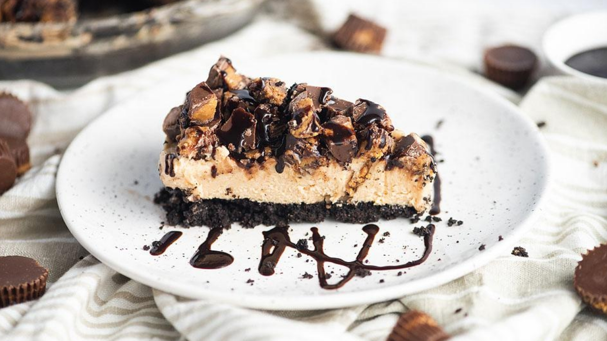 A slice of chocolate and peanut butter cheesecake with a dark cookie crust, topped with chopped chocolate pieces and chocolate drizzle, served on a white plate with additional chocolate candies nearby.