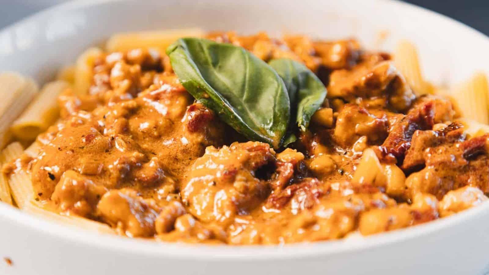 A close-up of rigatoni pasta topped with a creamy tomato-based sauce containing chunks of meat and garnished with a fresh basil leaf, served in a white bowl.