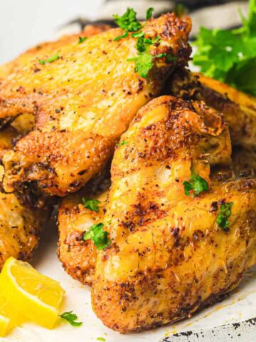 Close-up of a plate of seasoned and grilled chicken wings garnished with herbs. Lemon wedges are visible in the background, adding a hint of color. The wings appear crispy and well-cooked, resting on a dark plate.