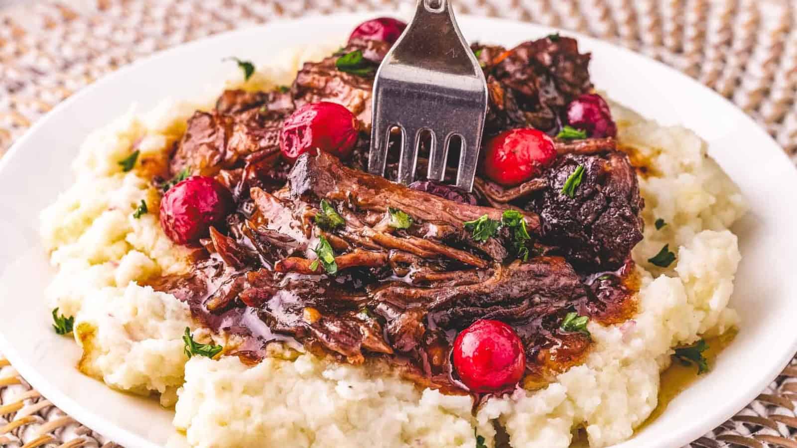 Shredded pot roast with gravy and cranberries on mashed potatoes, with a fork on a white plate.