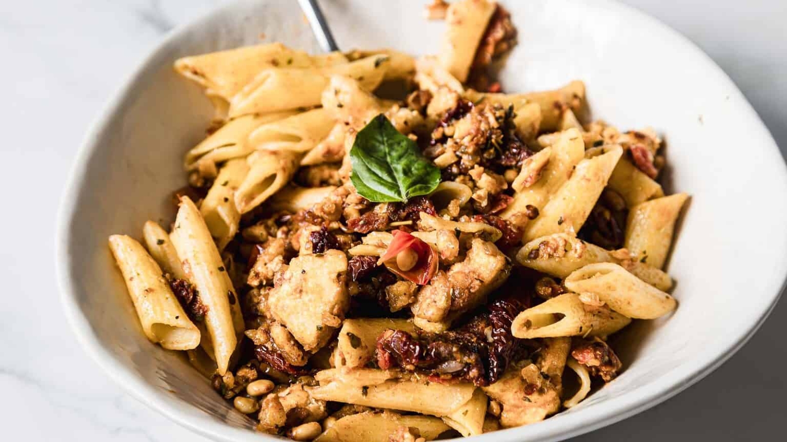 A white bowl filled with penne pasta mixed with chunks of chicken, sun-dried tomatoes, and garnished with fresh basil leaves. A fork is placed in the bowl, and a small dish is visible in the background.