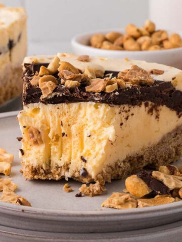A slice of cheesecake with a crumb crust and chocolate topping, garnished with chopped peanuts, served on a plate with additional peanuts, chocolate chips, and waffle cones nearby.