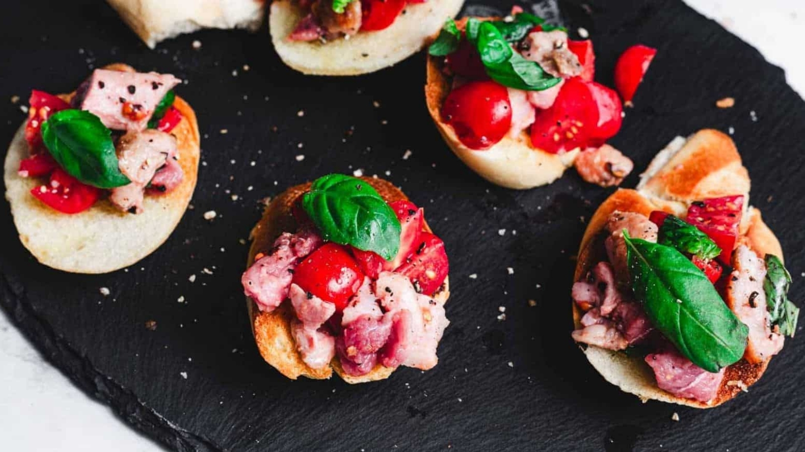 Slices of baguette topped with diced tomatoes, bacon, and basil leaves are arranged on a black slate surface. The ingredients are seasoned with cracked black pepper.