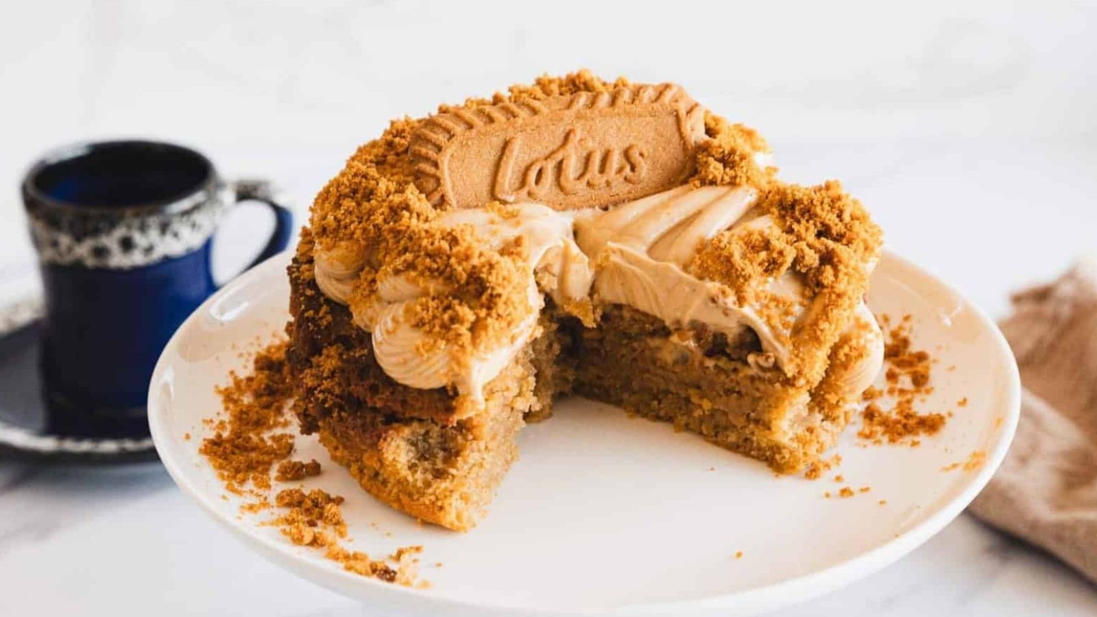 A partially sliced cake with light brown frosting and crumbled Lotus Biscoff cookies on top, displayed on a white cake stand. A blue cup sits in the background.