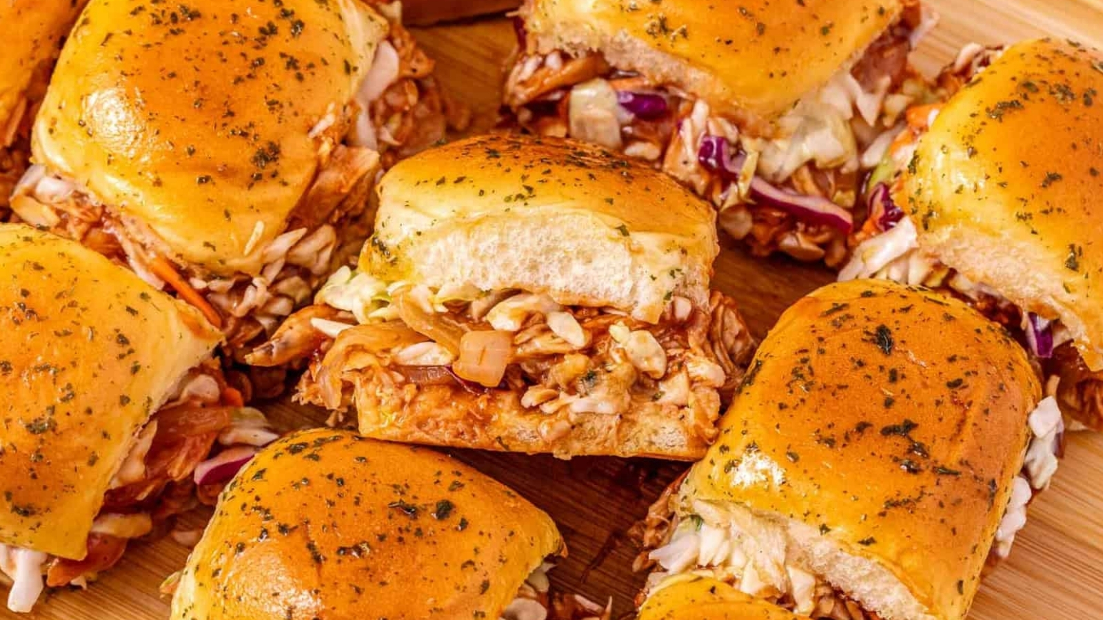 Several barbecue chicken sliders with coleslaw on golden buns are arranged on a wooden surface.
