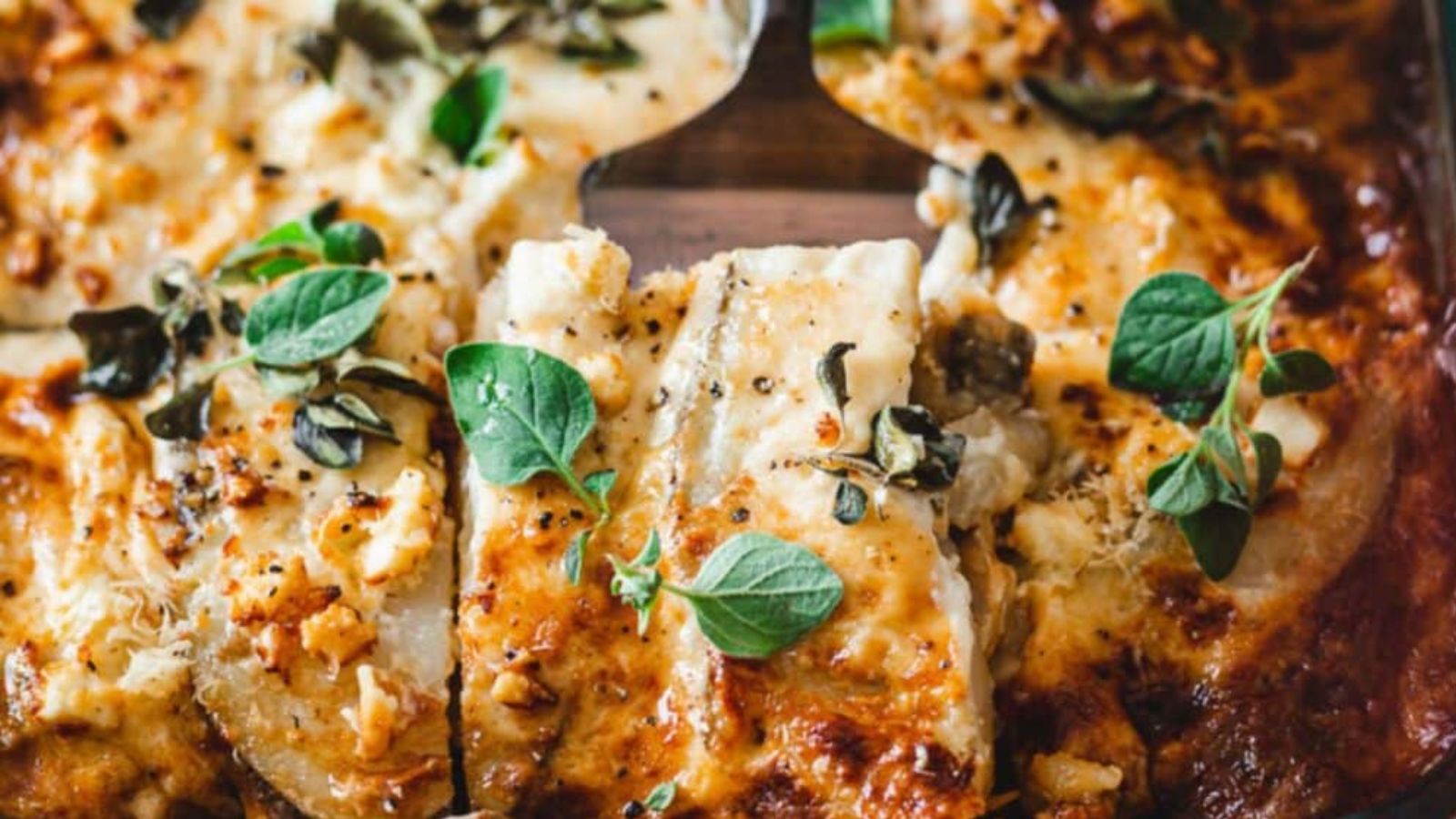 Close-up of a baked casserole dish featuring layers of cheese with crispy golden edges, topped with fresh green herbs.