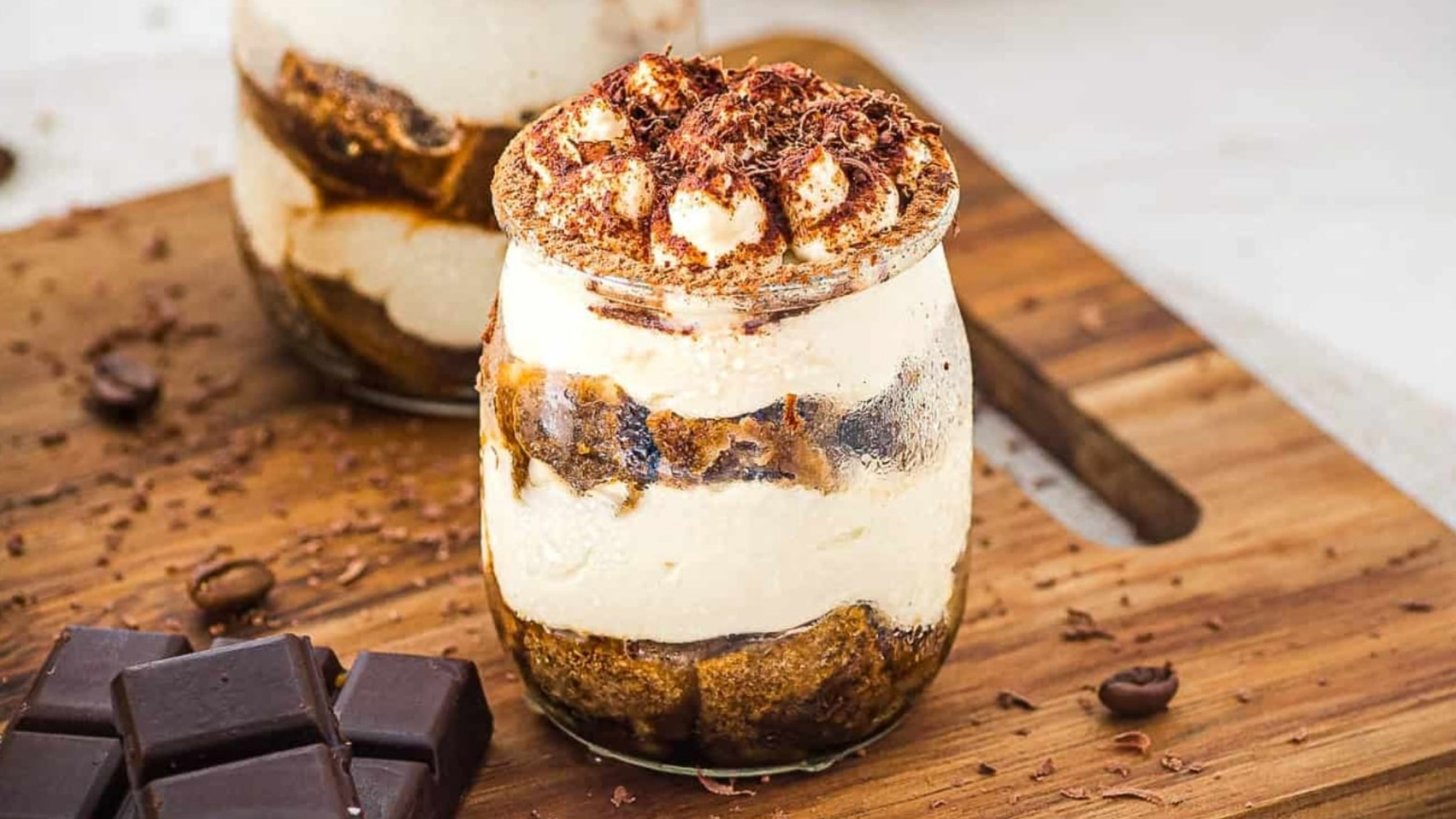 Two glass jars of tiramisu topped with cocoa and chocolate shavings, next to pieces of dark chocolate.