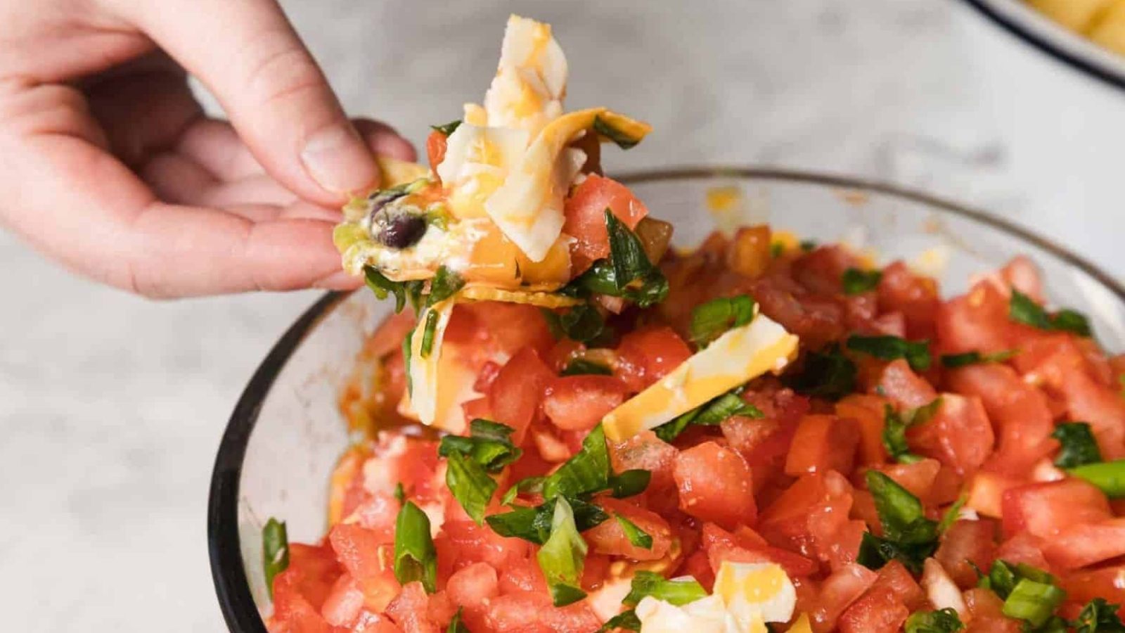 A person holds a chip topped with diced tomatoes, green onions, cheese, and olives, over a bowl filled with the same ingredients. The background is a light-colored countertop.