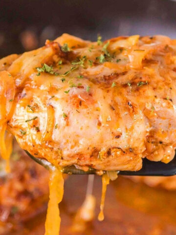 Spatula lifting a piece of chicken topped with melted cheese from a skillet with sauce visible below.