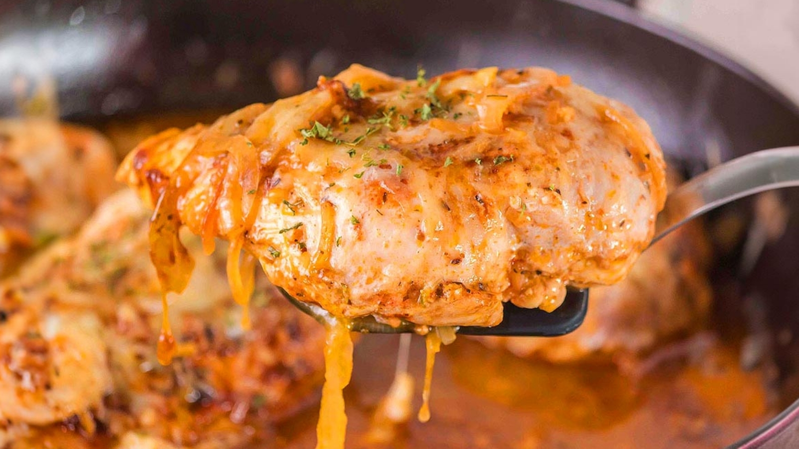 Spatula lifting a piece of chicken topped with melted cheese from a skillet with sauce visible below.