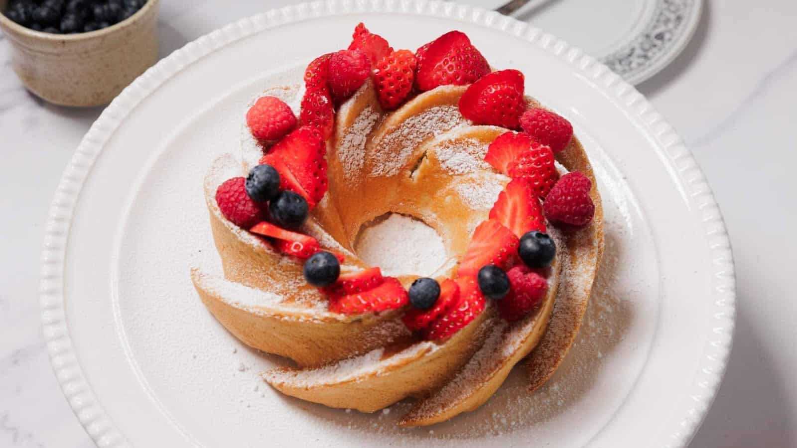 A Bundt cake topped with sliced strawberries, whole blueberries, and a dusting of powdered sugar sits on a white plate. A bowl of blueberries and a decorative plate are visible in the background.