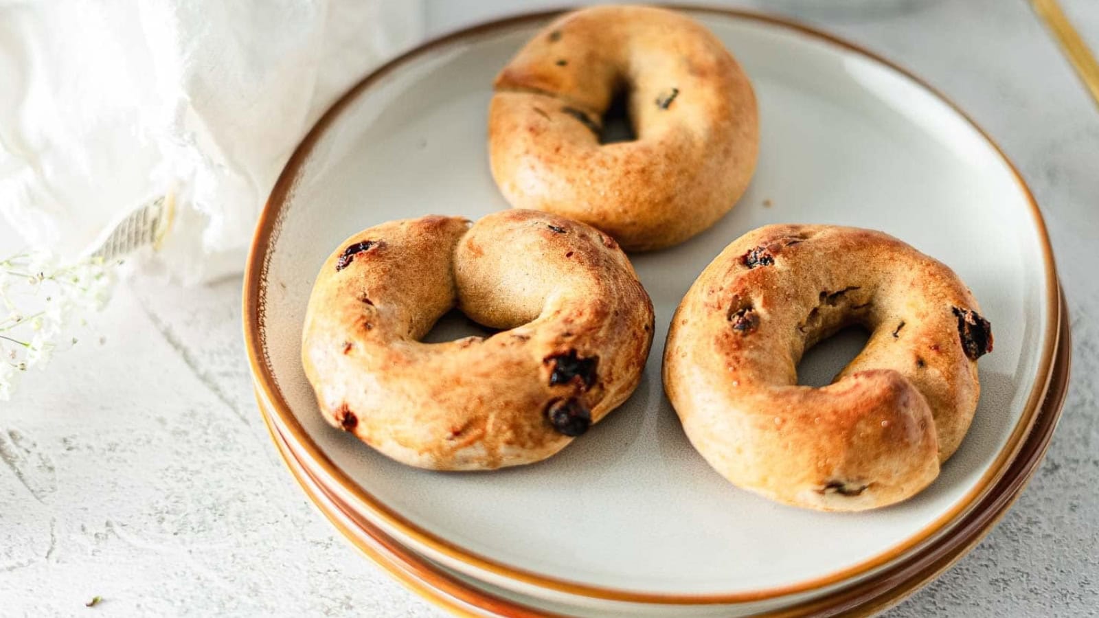 Three homemade bagels with raisins on a white plate, set on a light-colored table.