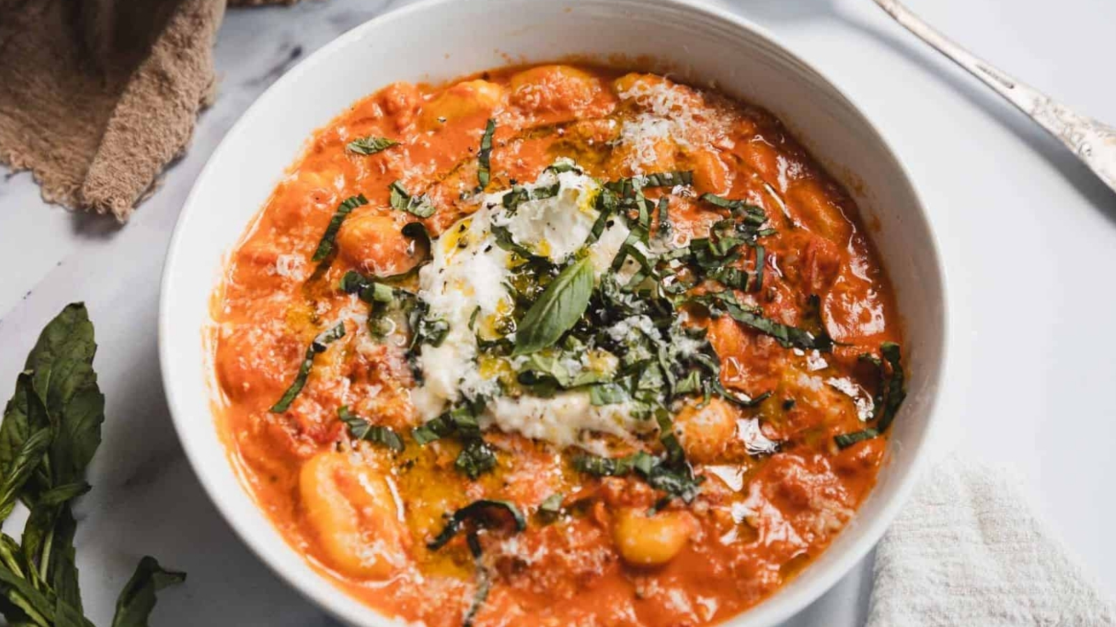 A white bowl filled with tomato-based gnocchi topped with creamy cheese, chopped fresh basil, and a drizzle of olive oil sits on a white surface with a fork, napkin, and basil leaves nearby.
