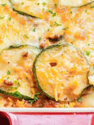 A baked dish in a red casserole dish featuring layers of melted cheese, zucchini slices, and a seasoned meat mixture. The surface is lightly browned, garnished with chopped herbs.